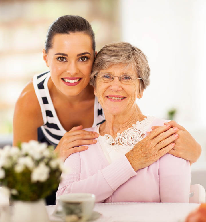 Adult daughter hugging Mom from behind at a table, both smiling